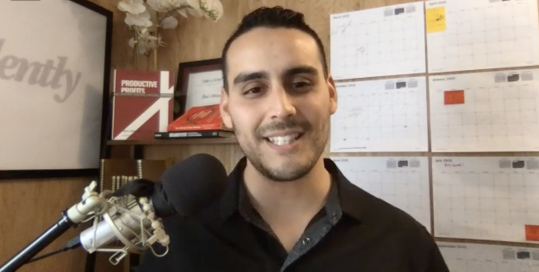 A person smiles in front of a microphone, books, and calendars on a wall. The setting appears to be a home office.